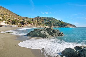 A coastal beach with rocky formations, clear blue water, and a hillside with buildings.