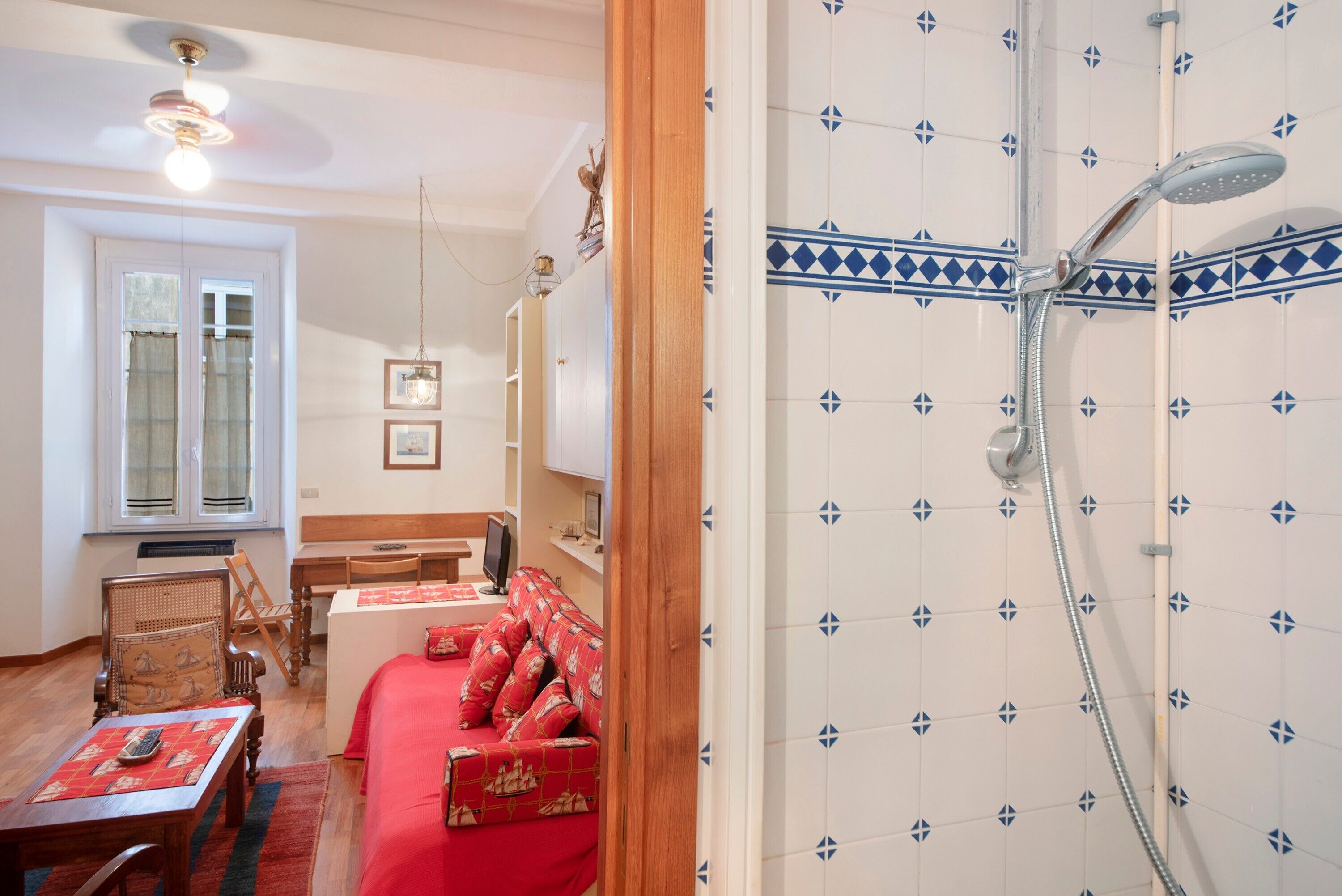 A room with a wooden floor, a red sofa, a dining table, a TV, and a shower with blue diamond-patterned tiles.