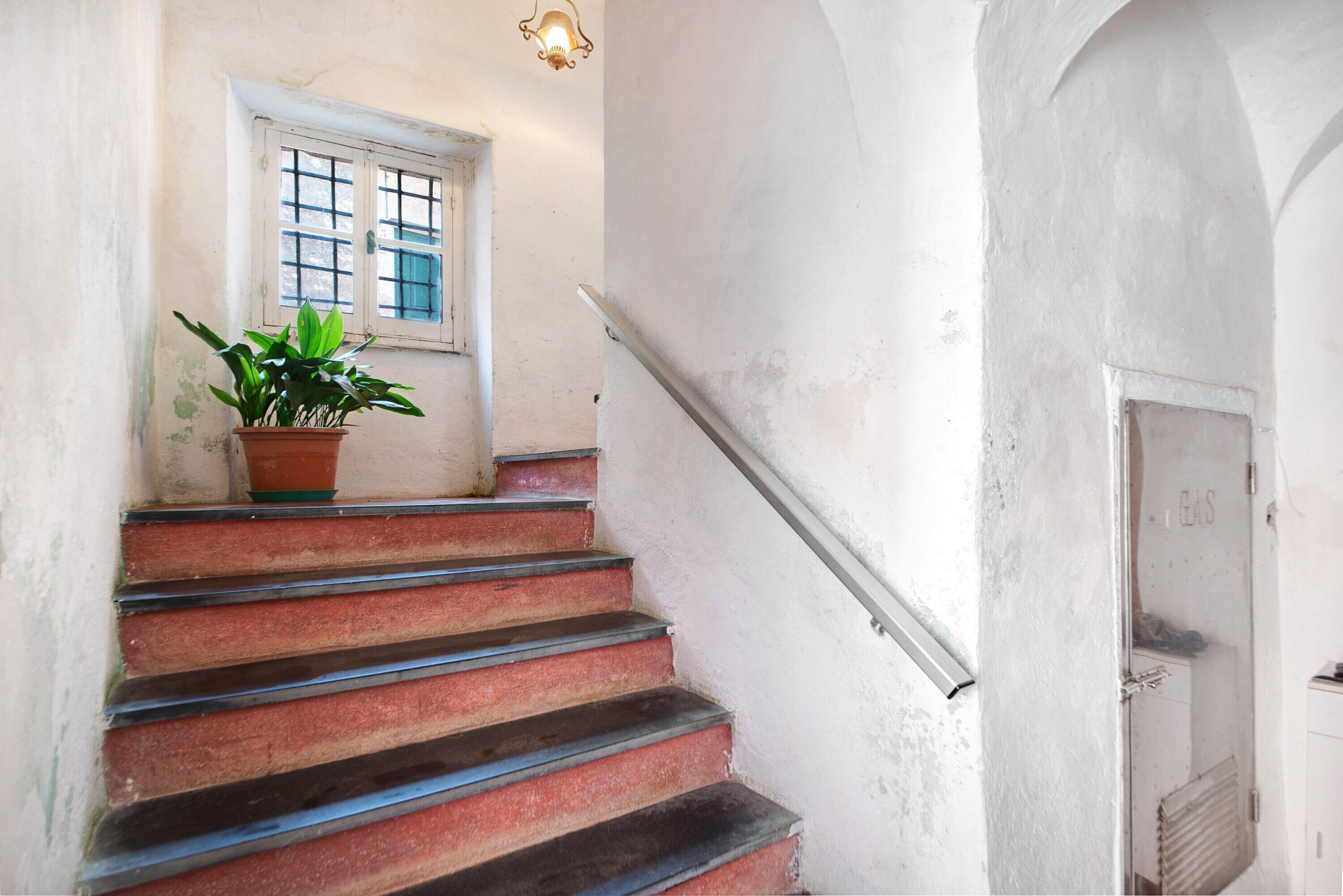 Eine Treppe mit einem Blumentopf, ein Fenster mit Gittermuster und eine Tür mit der Aufschrift 'E GAS'.