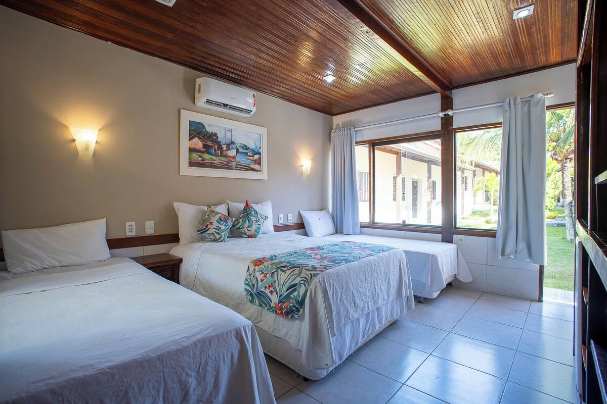 A hotel room with two beds, a ceiling fan, an air conditioning unit, a painting on the wall, and a window with curtains.