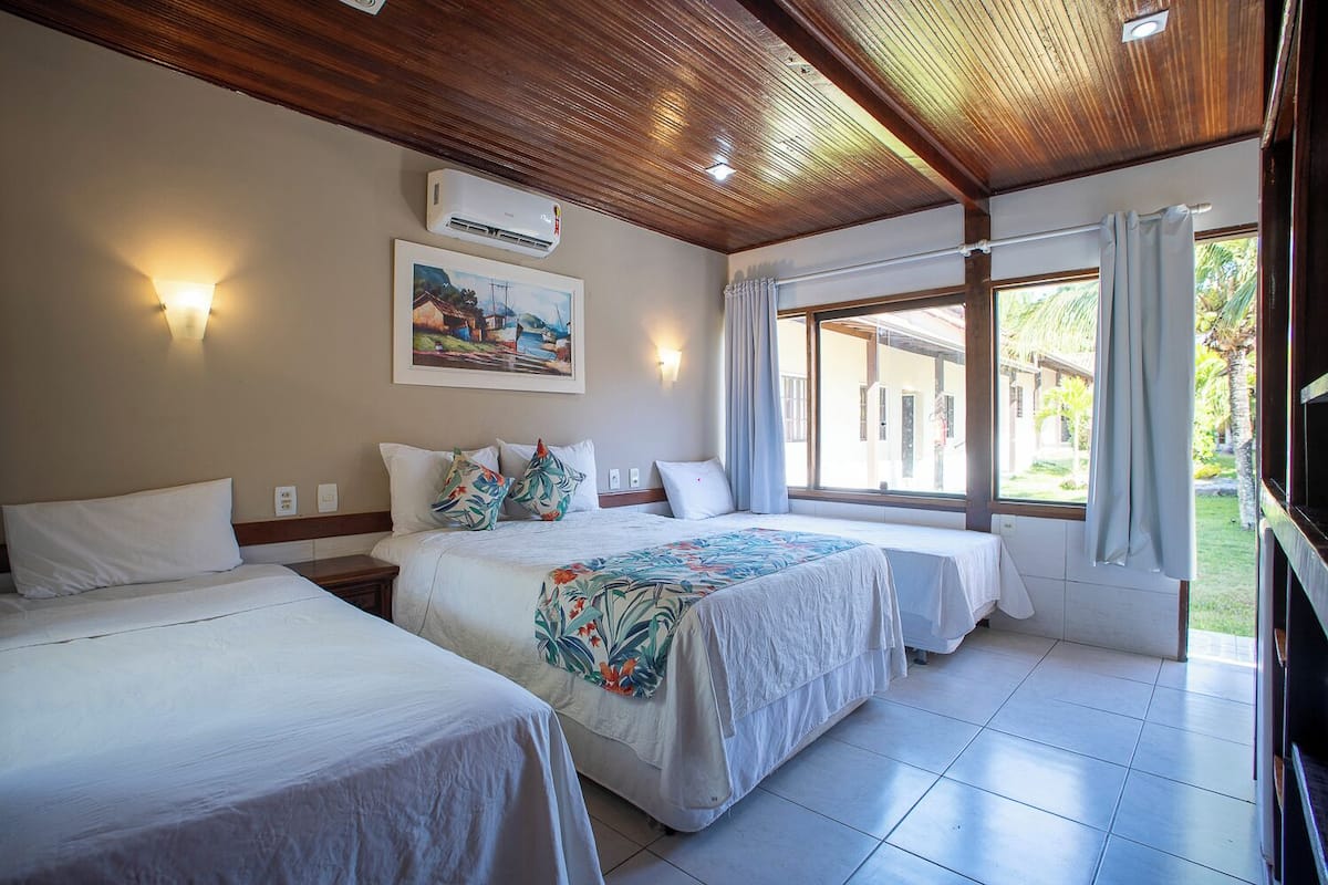 A hotel room with two beds, a ceiling fan, an air conditioning unit, a painting on the wall, and a window with curtains.