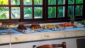 A buffet table with various dishes, a wooden chair in the foreground, and a window with a view of greenery. - Pousada Maria Maria Buzios (Búzios)