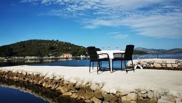 A table and chairs by a lake with a stone wall and a hill in the background. - Apartment Pascal (Janjina)