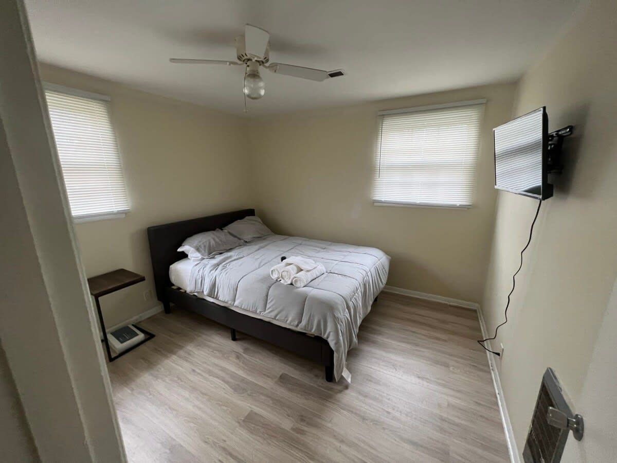 A bedroom with a bed, a ceiling fan, a wall-mounted TV, and a window with blinds.