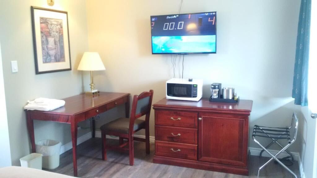 Habitación de hotel con un escritorio de madera, microondas, un mueble pequeño, una silla, una lámpara, un cuadro en la pared y un televisor que muestra un reloj digital.