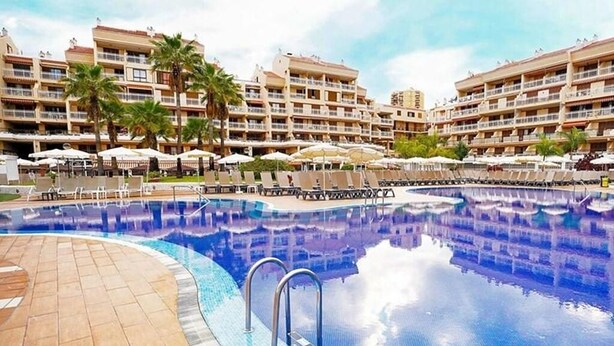A hotel pool area with multiple lounge chairs and umbrellas, surrounded by multi-story buildings with balconies.