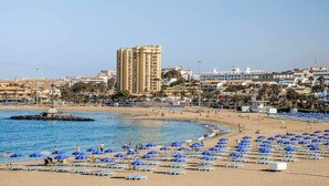 A beach with numerous blue umbrellas and sun loungers, a large building in the background, and a clear blue sky.