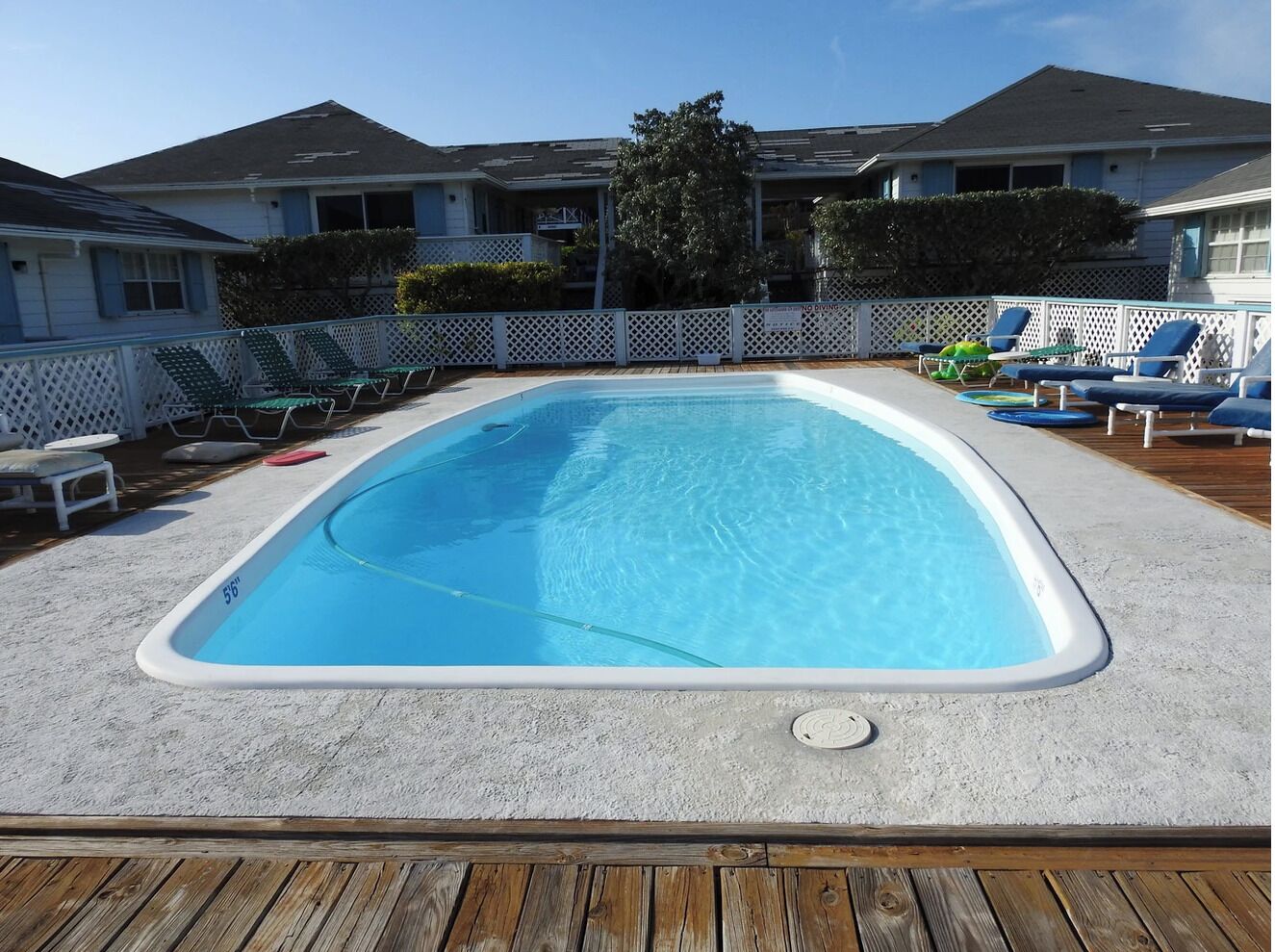 A rectangular above-ground pool with a white deck surrounding it, a wooden deck in the foreground, and a white fence with lounge chairs on one side.