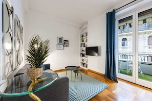 A modern living room with a glass coffee table, a sofa, a bookshelf, and a TV mounted on the wall.