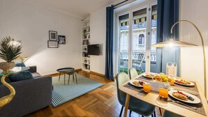 Ein modernes Wohnzimmer mit einer Couch, einem Esstisch gedeckt für das Frühstück, einem Bücherregal und Blick auf die Stadt durch große Fenster.