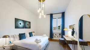 Ein modernes Hotelzimmer mit einem großen Bett, Nachttischen, einem Schreibtisch und einem großen Spiegel. An den Wänden sind abstrakte Kunstwerke angebracht und durch das Fenster ist die Stadt zu sehen.
