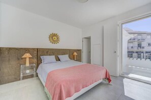 Ein Schlafzimmer mit einem Bett, zwei Nachttischlampen, einer Wanduhr und Blick auf Gebäude durch ein großes Fenster.