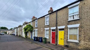 A row of terraced houses with distinctively colored doors, a clear road, and a cloudy sky. - Bright Beautiful Modern 2 Bed Home Pass The Keys (Cambridge)