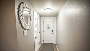Um corredor estreito com uma porta branca, um espelho decorativo e um tapete de entrada.