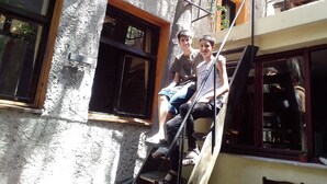 Two individuals seated on a wooden staircase outside a building with a rough-textured exterior and multiple windows. - Titan House Hostel (Izmir)