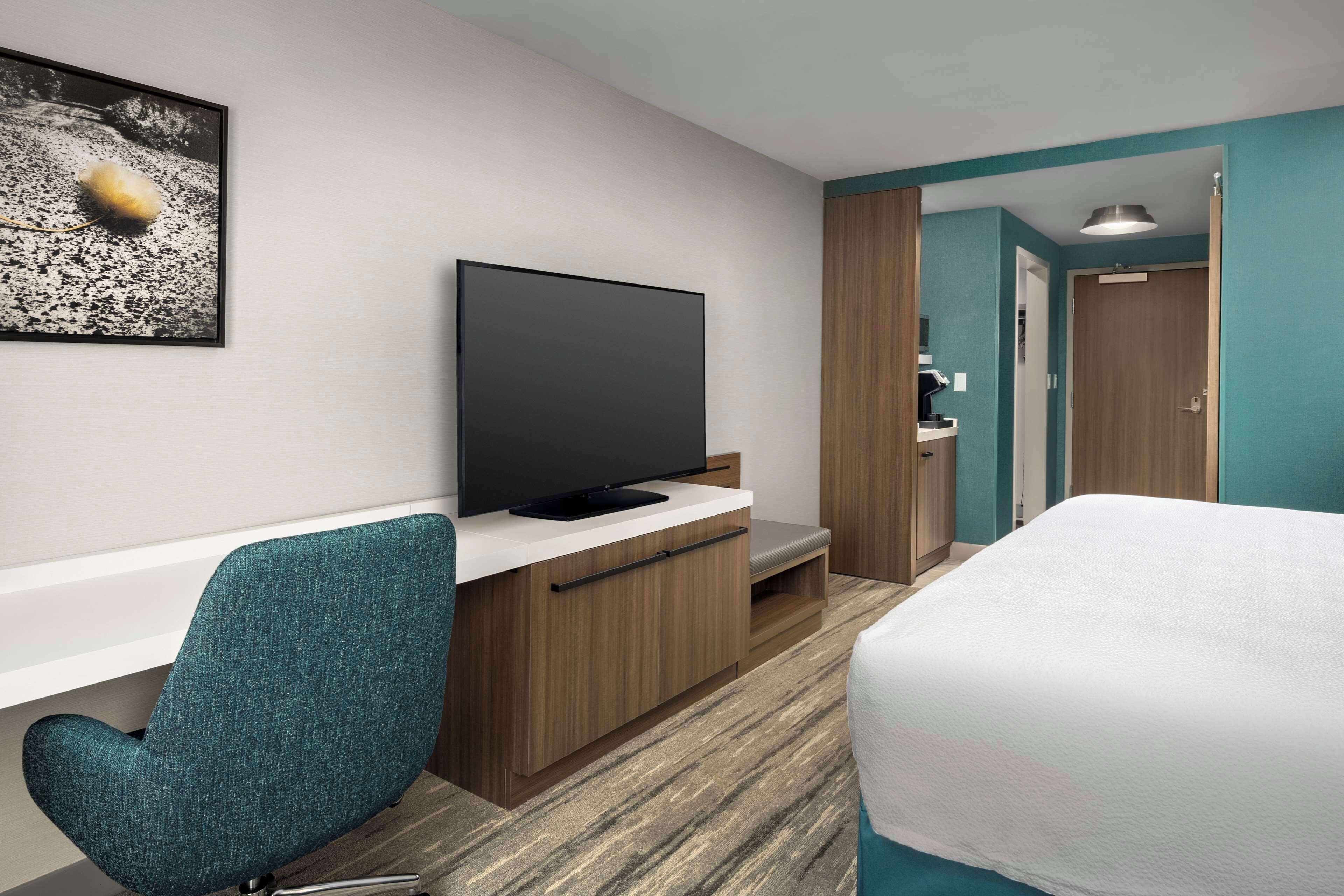 Una habitación de hotel con una televisión de pantalla plana sobre un mueble de madera, una silla color turquesa, una cama con ropa blanca y una puerta que da a un baño.