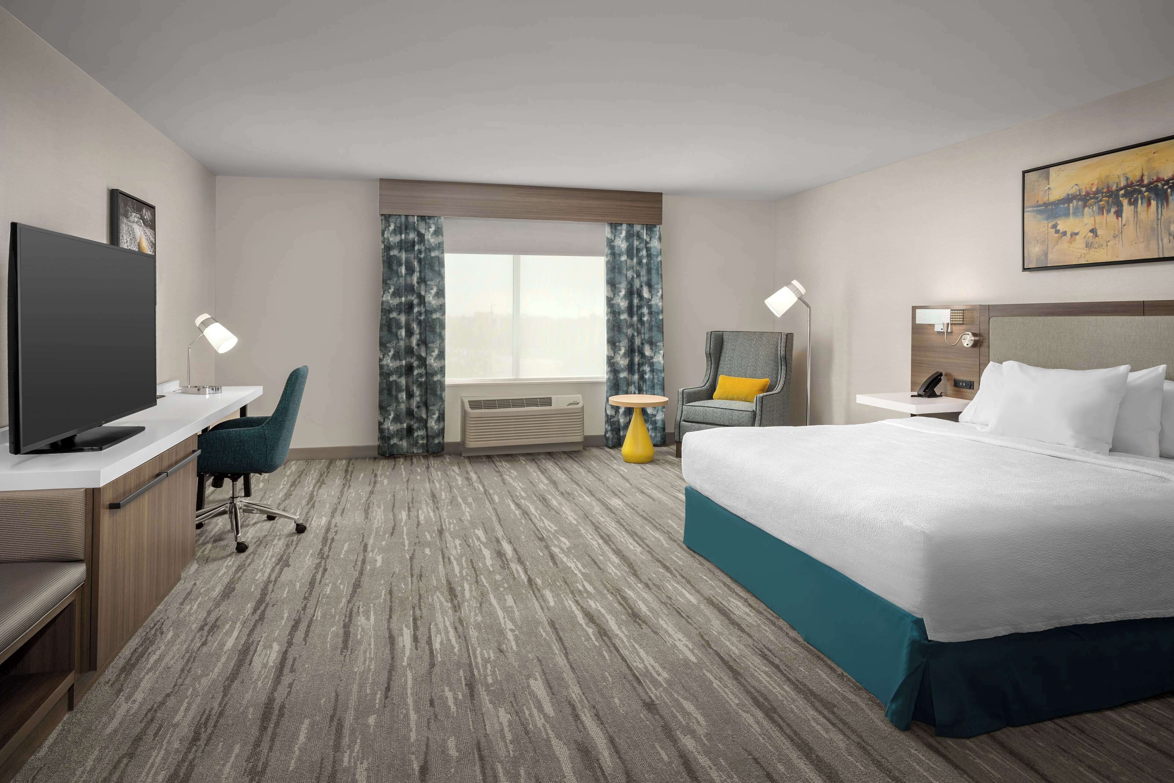 Une chambre d’hôtel avec un lit, un bureau avec une télévision, une chaise, une fenêtre avec des rideaux et un tableau au mur.