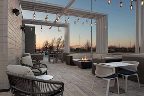 Een terras buiten met rieten meubels, een vuurkorf en hangende lampen.