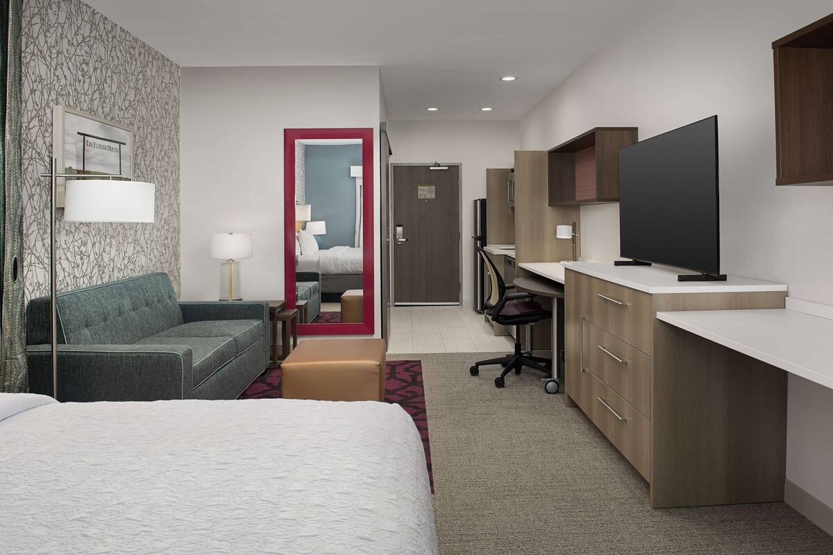 A hotel room with a bed, a sofa, a desk, a TV, and a mirror.