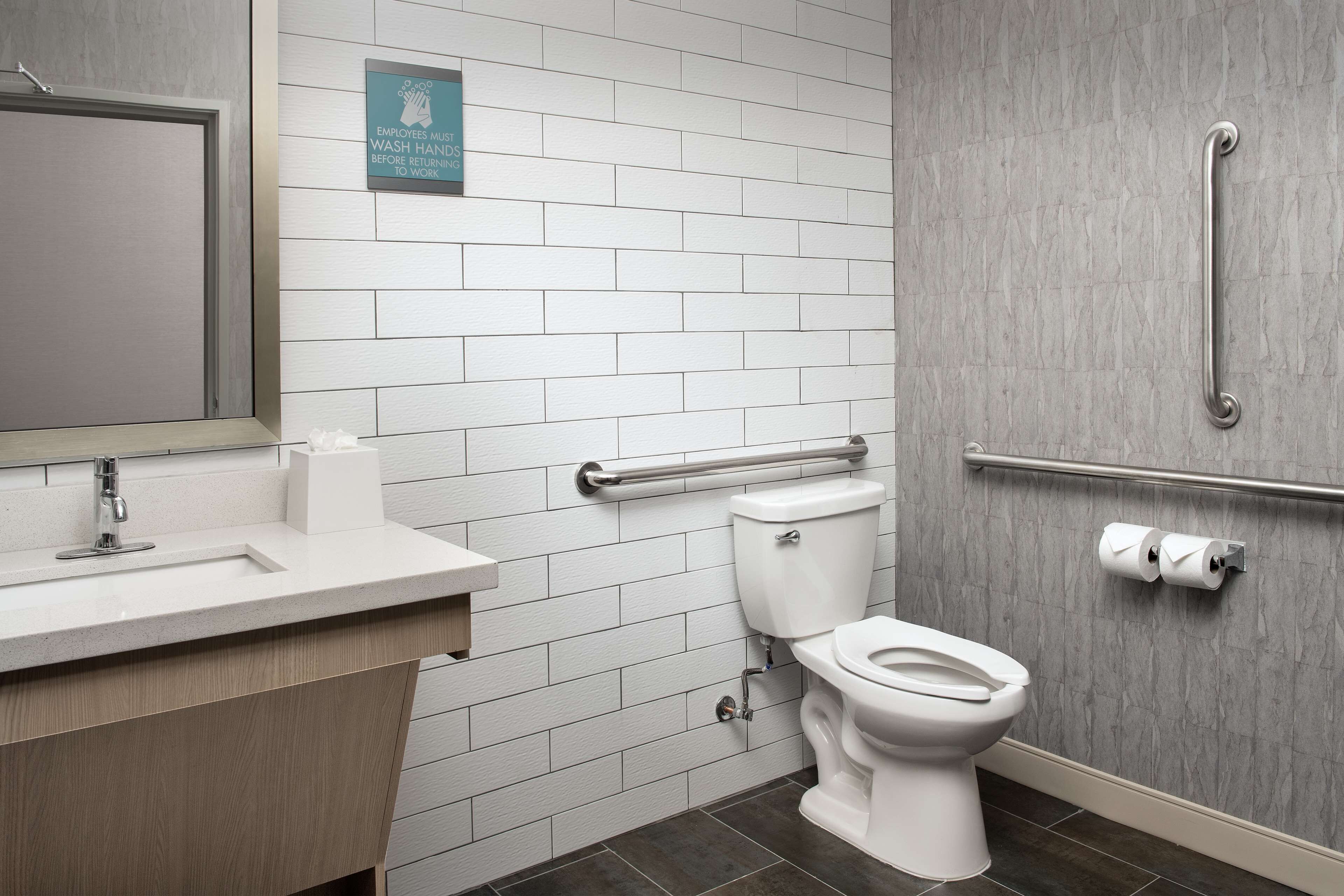 A modern bathroom with a white tiled wall, a sink with a mirror, a toilet with a grab bar, and a hand dryer.