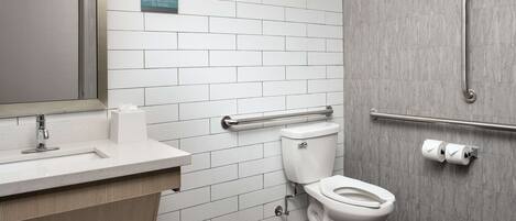 Une salle de bain moderne avec un mur recouvert de carreaux blancs, un lavabo avec un miroir, des toilettes équipées d’une barre d’appui et un sèche-mains.