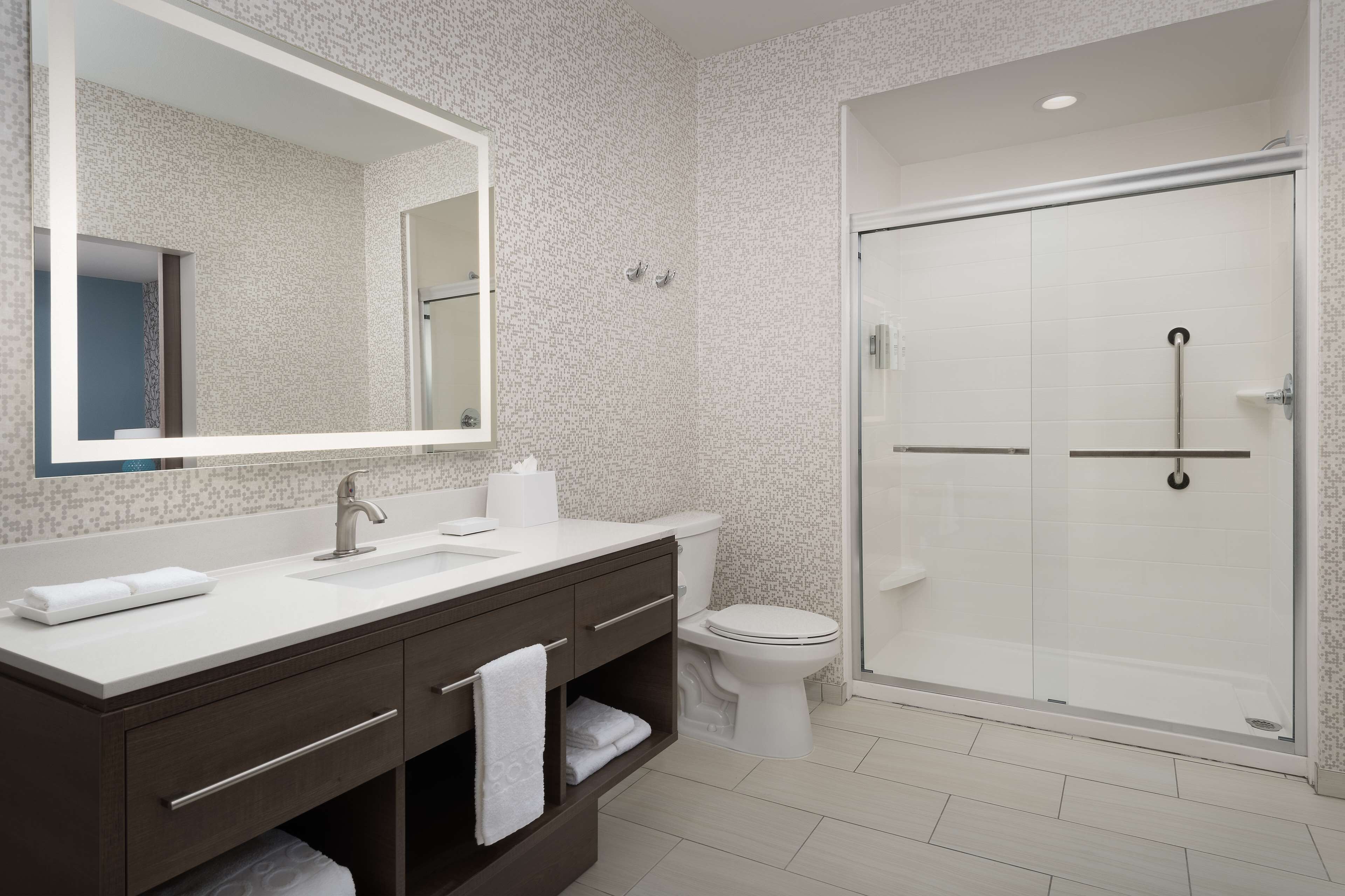 Un baño moderno con un espejo grande, un lavabo blanco, un mueble de tocador oscuro, un inodoro y un box de ducha de vidrio.