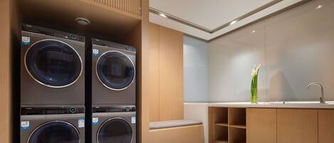 A modern laundry room with two stacked front-loading washers and dryers, wooden cabinetry, a built-in bench, and a sink with a vase of flowers.