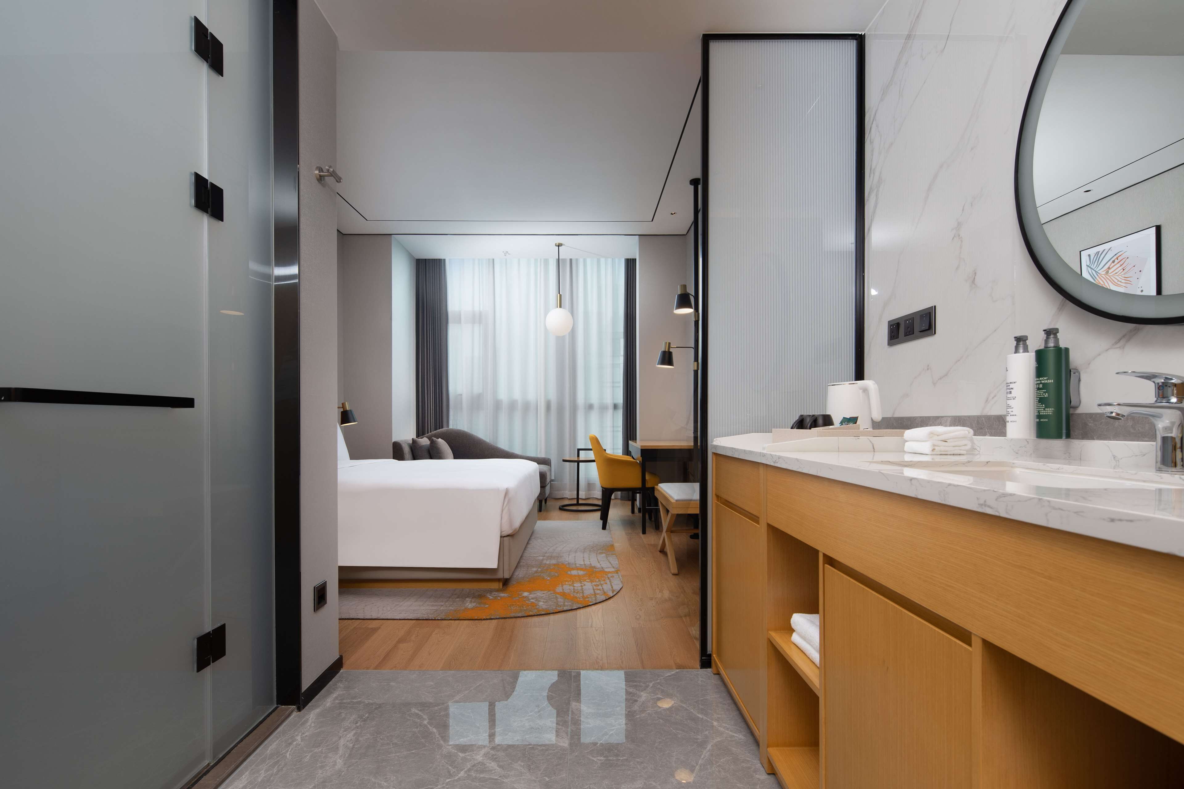 A modern hotel room with a bed, a desk, a chair, a bathroom with a sink and mirror, and a view of the city through large windows.