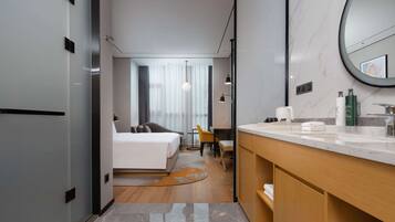 A modern hotel room with a bed, a desk, a chair, a bathroom with a sink and mirror, and a view of the city through large windows.
