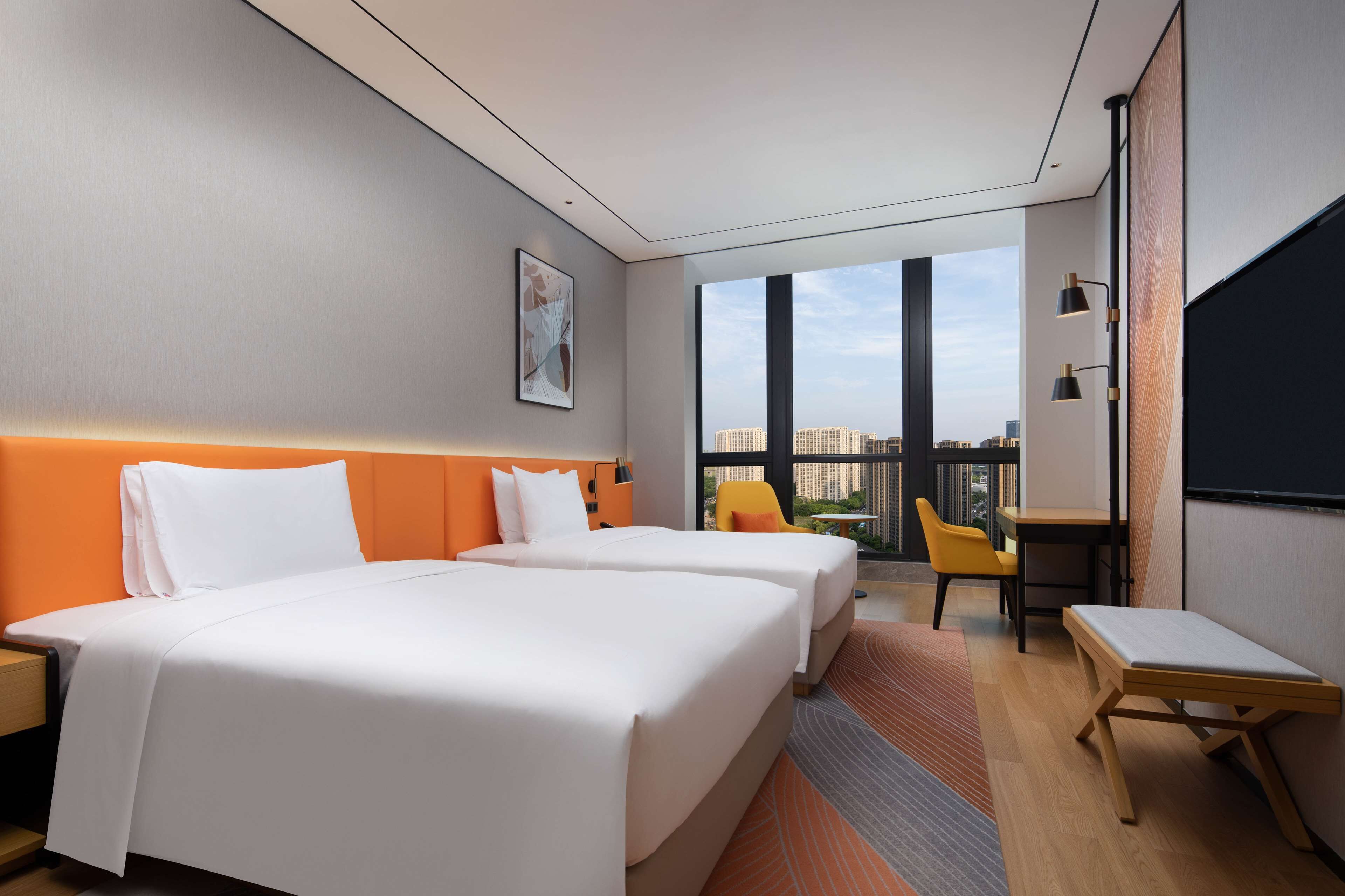 Ein Hotelzimmer mit zwei Betten, einem Schreibtisch, einem Stuhl und einem großen Fenster mit Blick auf die Stadt.
