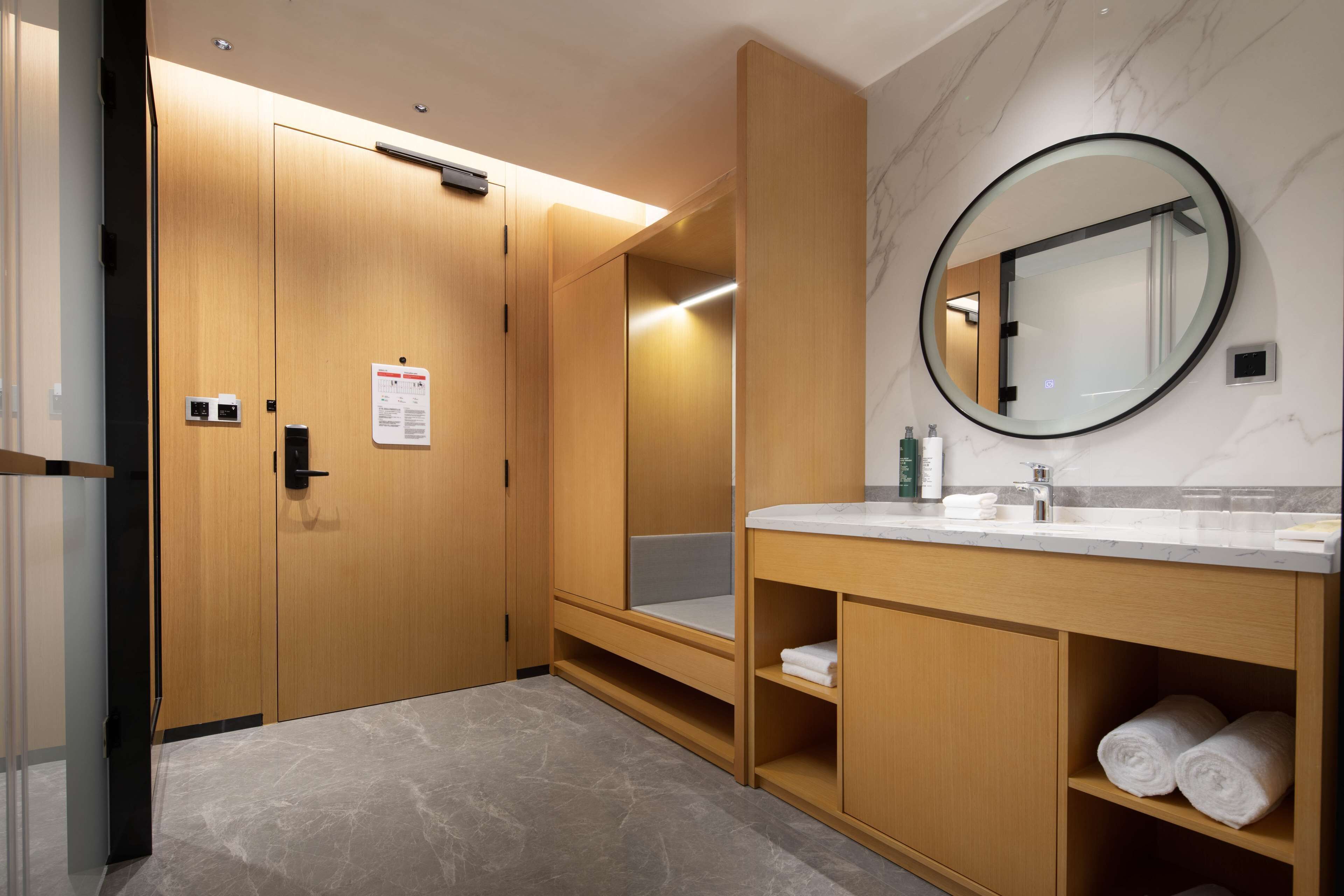 A modern bathroom with wooden cabinets, a marble floor, a round mirror, and a white countertop.