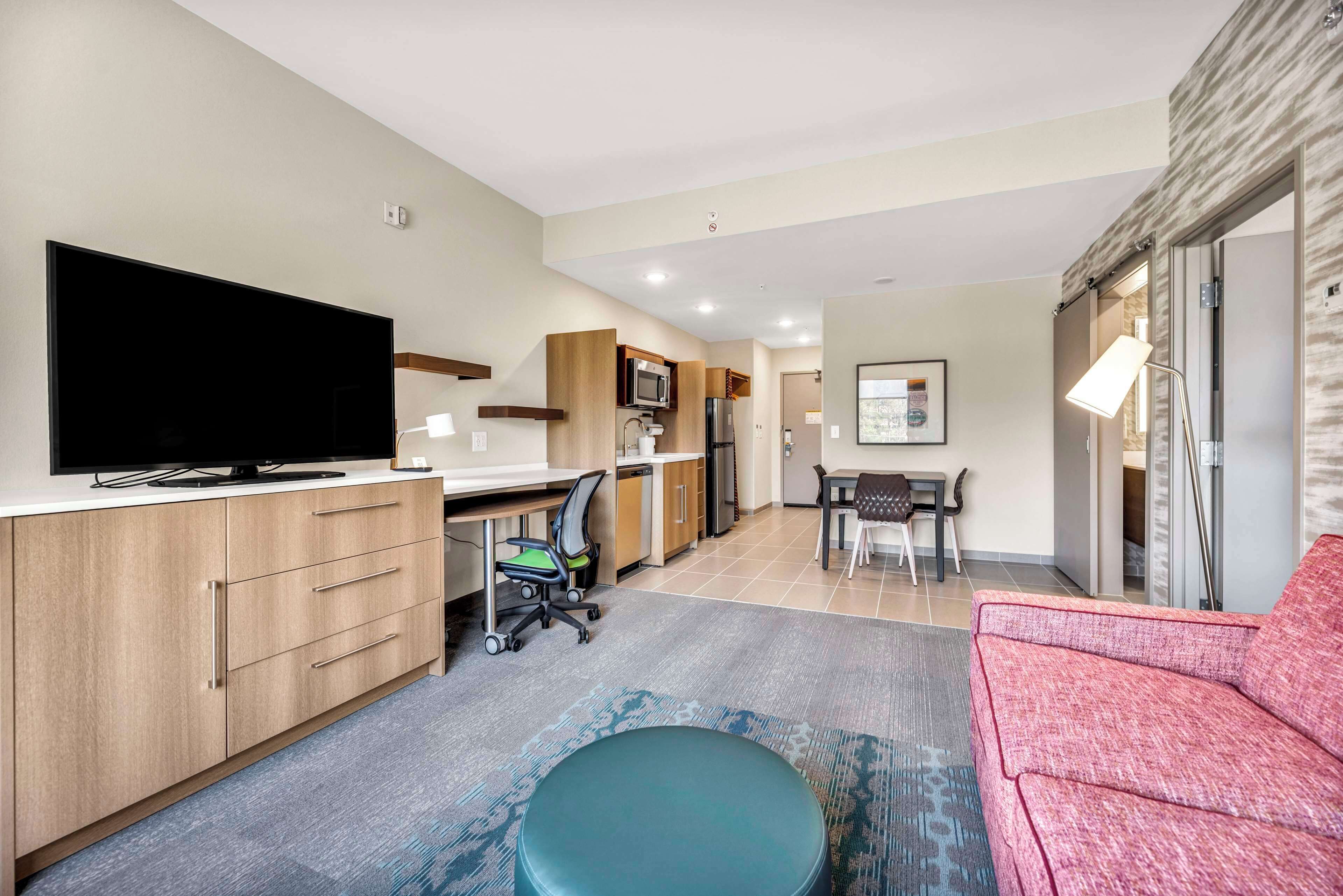 A modern hotel room with a flat-screen TV, wooden cabinets, a kitchenette, a dining area, and a red sofa.