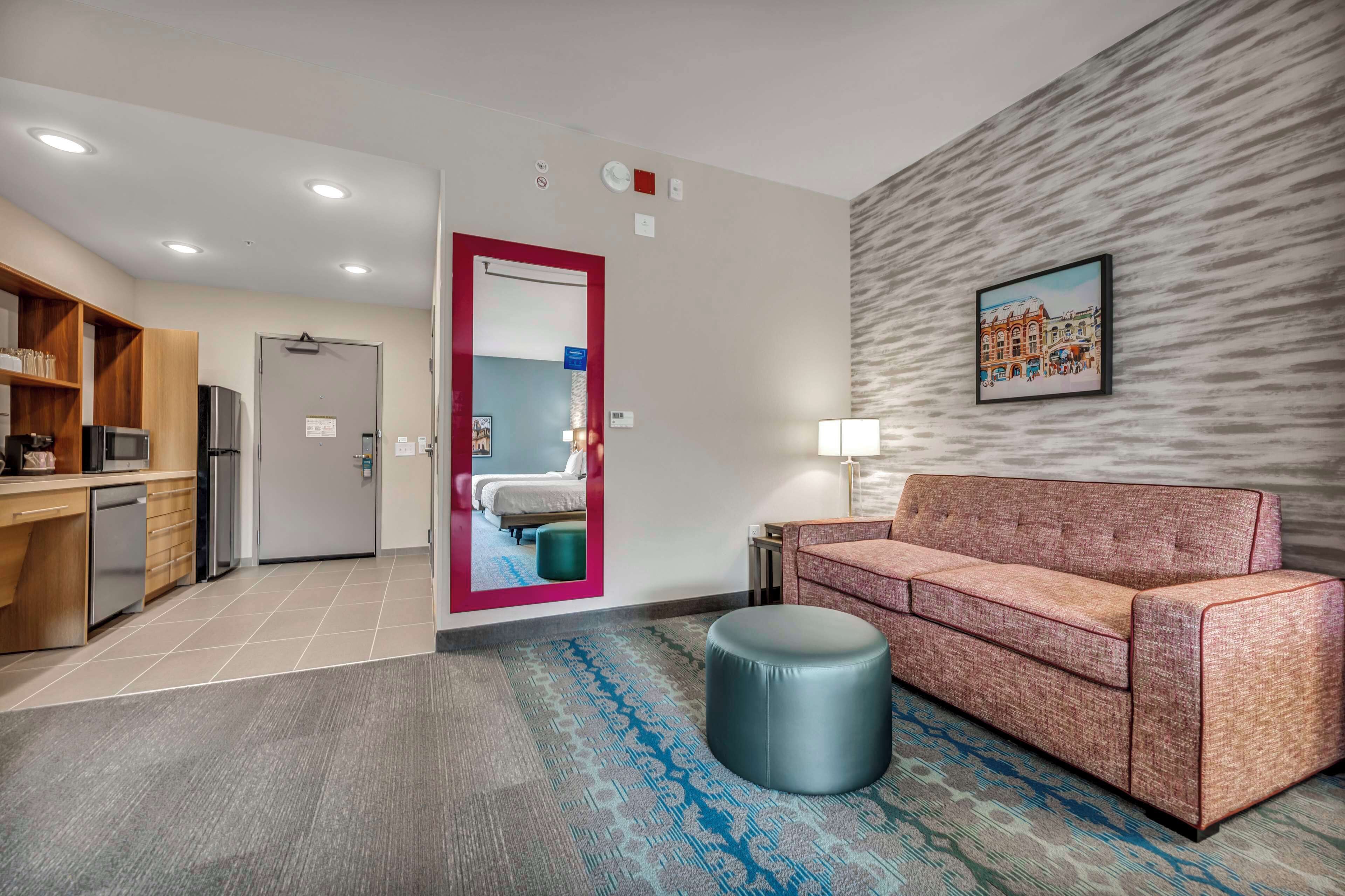 A hotel lobby with a kitchenette, a sofa, a round ottoman, a mirror, and a framed picture on the wall.