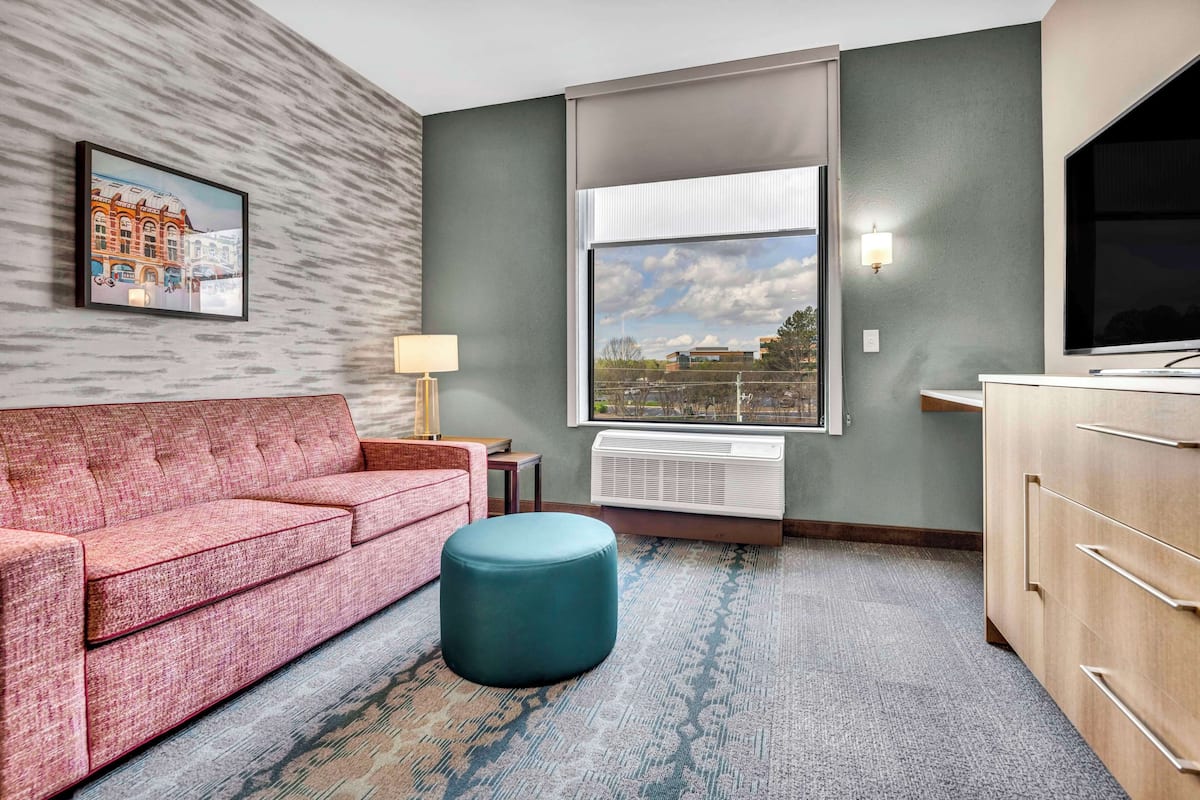 Een hotelkamer met een rode bank, een blauwgroene poef, een groot raam met uitzicht op de stad, een wandtelevisie en een ingelijste foto aan de muur.
