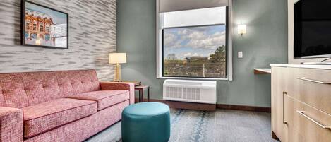 A hotel room with a red sofa, a teal ottoman, a large window with a city view, a wall-mounted TV, and a framed picture on the wall.