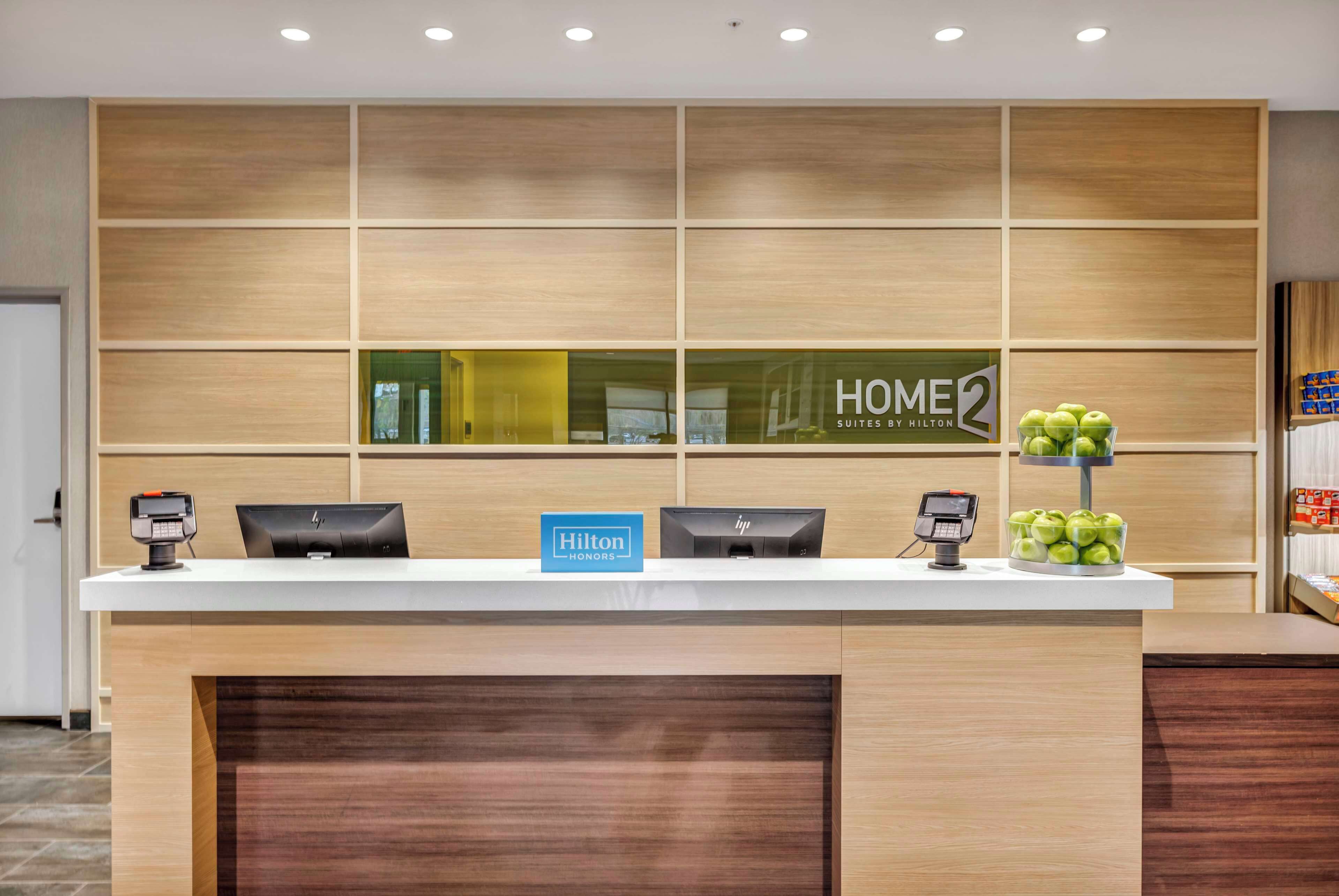 Un mostrador de recepción de hotel con diseño de paneles de madera, un letrero que dice 'HOMER 2 Suites by Hilton' y una exhibición de frutas.
