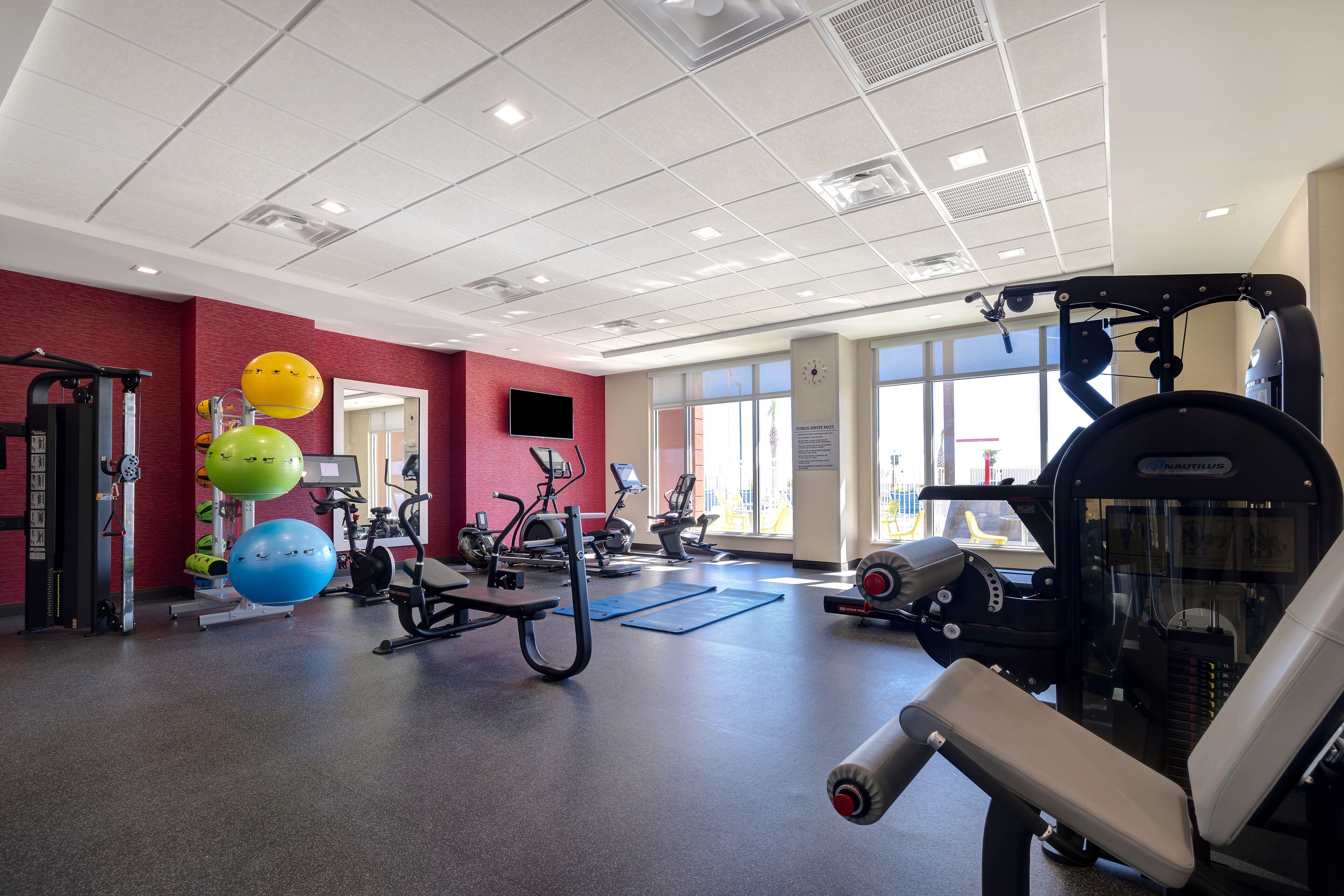A gym with various exercise equipment, a red wall, and large windows.