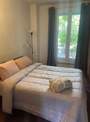 A neatly made bed with a folded towel, a standing floor lamp, and a window with curtains.