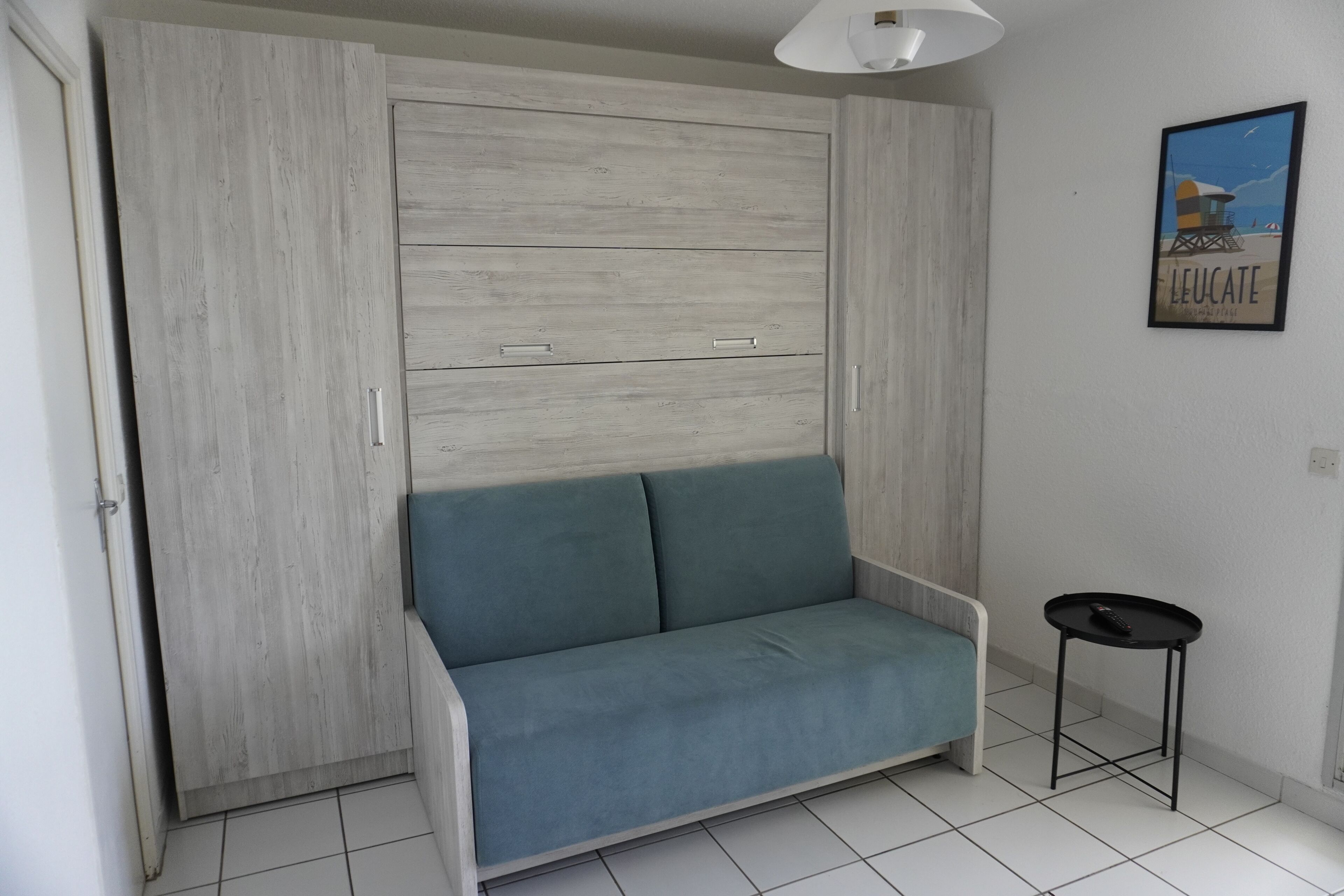 A room with a light wooden wardrobe, a teal sofa, a small black table, and a framed picture on the wall.