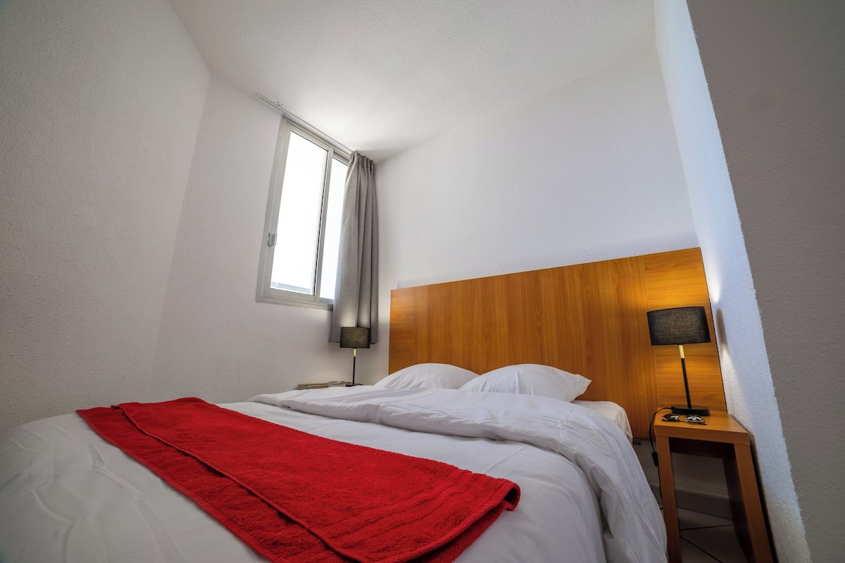 Een hotelkamer met een bed, een rode deken, een houten hoofdeinde, een nachtkastje met een lamp en een raam met gordijnen.