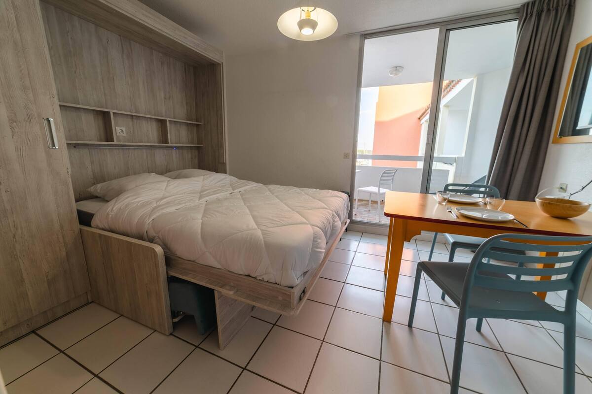 Een compacte kamer met een uitschuifbaar bed, een eettafel met stoelen en een glazen schuifdeur naar een buitenruimte.
