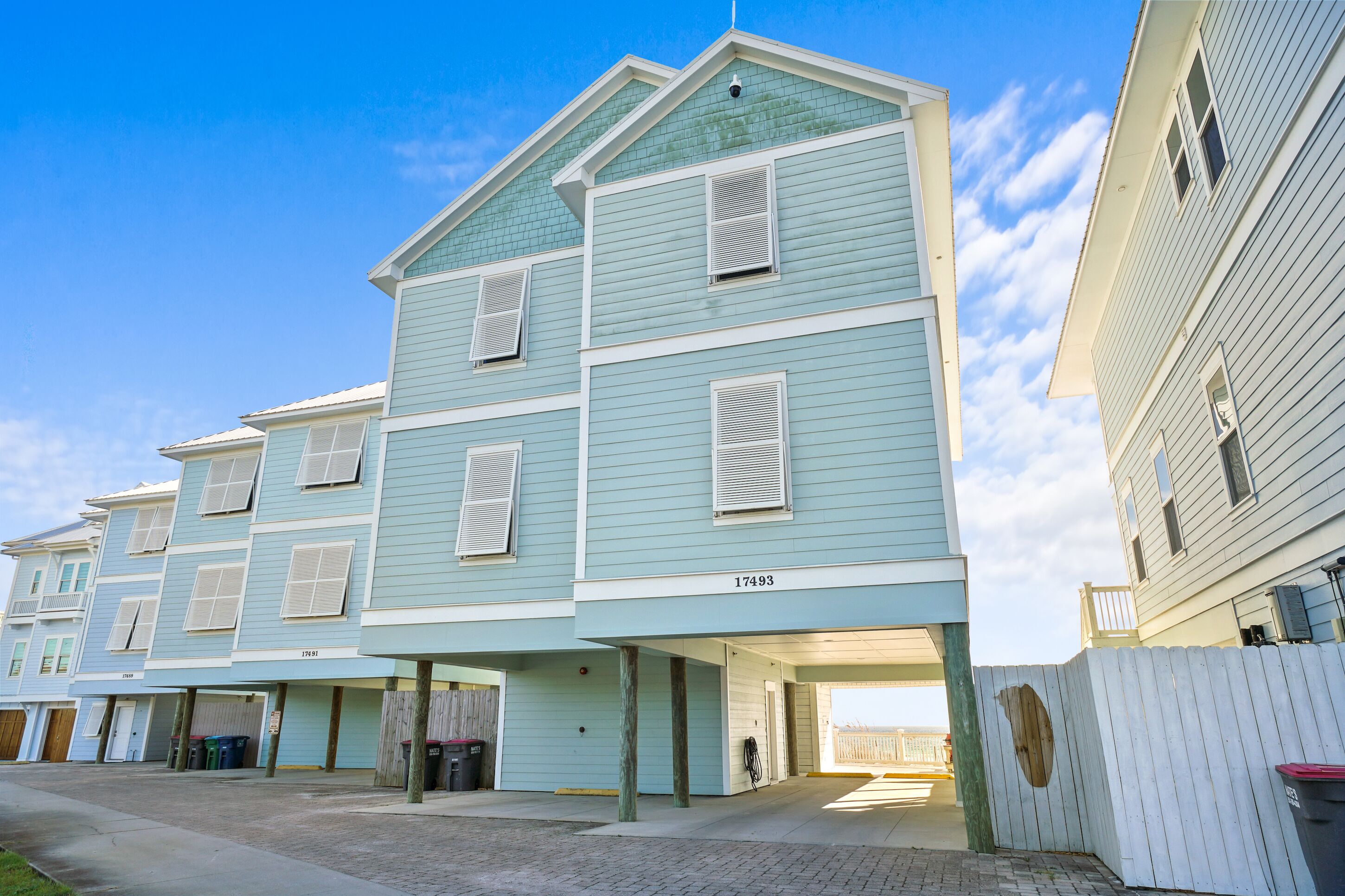 A multi-story residential building with a light blue exterior, numbered 17493, featuring a garage entrance and a visible wooden fence.