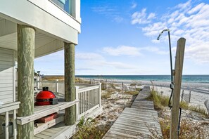 En träbrygga som leder till en strand, en röd grill på en veranda och ett trästaket längs strandkanten.