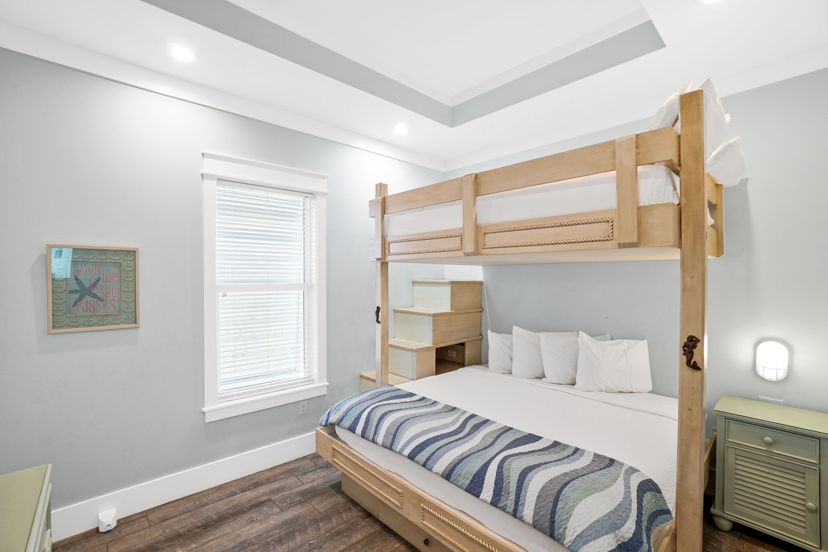 A bunk bed room with a wooden headboard, white bedding, a small bedside table, and a window with blinds.