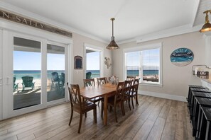 En matplats med ett stort träbord och stolar, en hänglampa och utsikt över stranden genom stora glasdörrar.