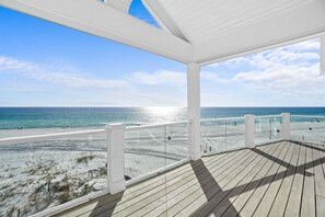 Une terrasse en bois dotée d’une balustrade en verre, offrant une vue sur une plage où le ciel est dégagé et la mer est calme.