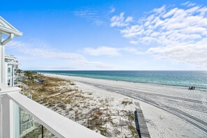 Strandudsigt fra en balkon med klart himmel, sandstrand og en bygning med hvid facade.