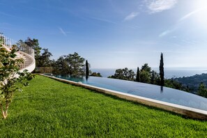A curved infinity pool with a clear view of the ocean, surrounded by well-maintained grass and tall trees.
