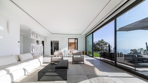 Una sala de estar moderna con sofá, mesa de centro y sillas, ventanas amplias con vistas, y una estantería.