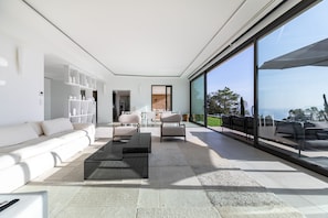 A modern living room with a sofa, coffee table, and chairs, large windows with a view, and a bookshelf.
